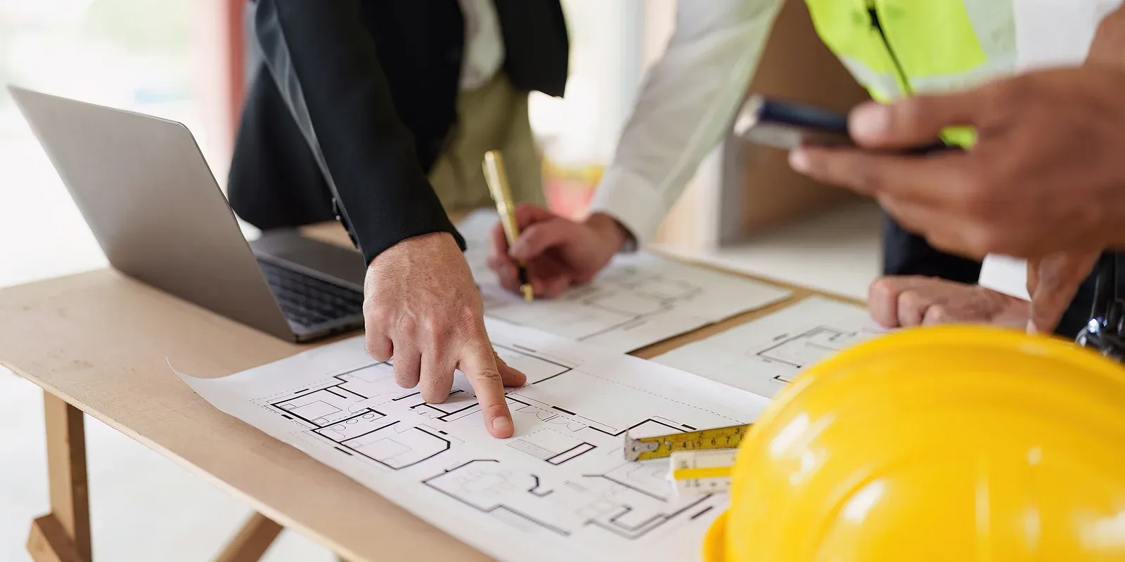People stand and gesture at a floorplan on a table with a laptop to the left and a yellow hard hat to the right.