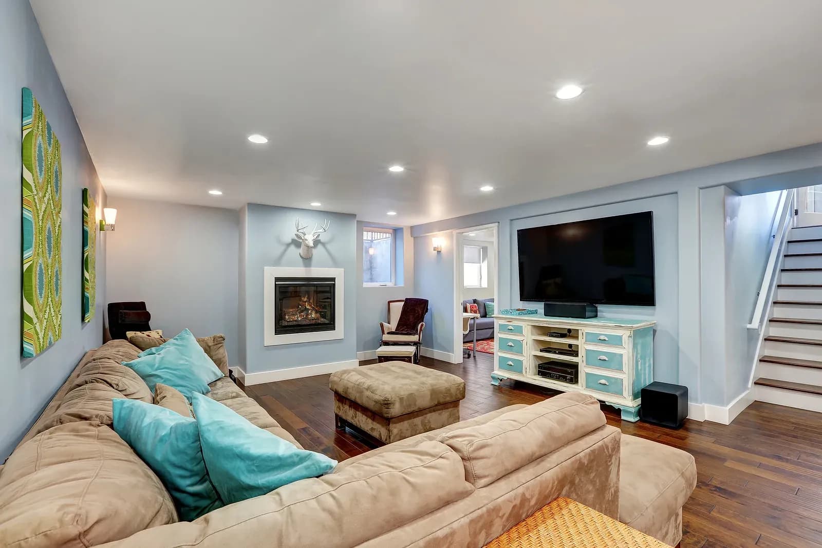 A photo of a finished basement with brown wood floors, blue painted walls, a sectional couch. A TV is mounted on the wall above a console table to the right.