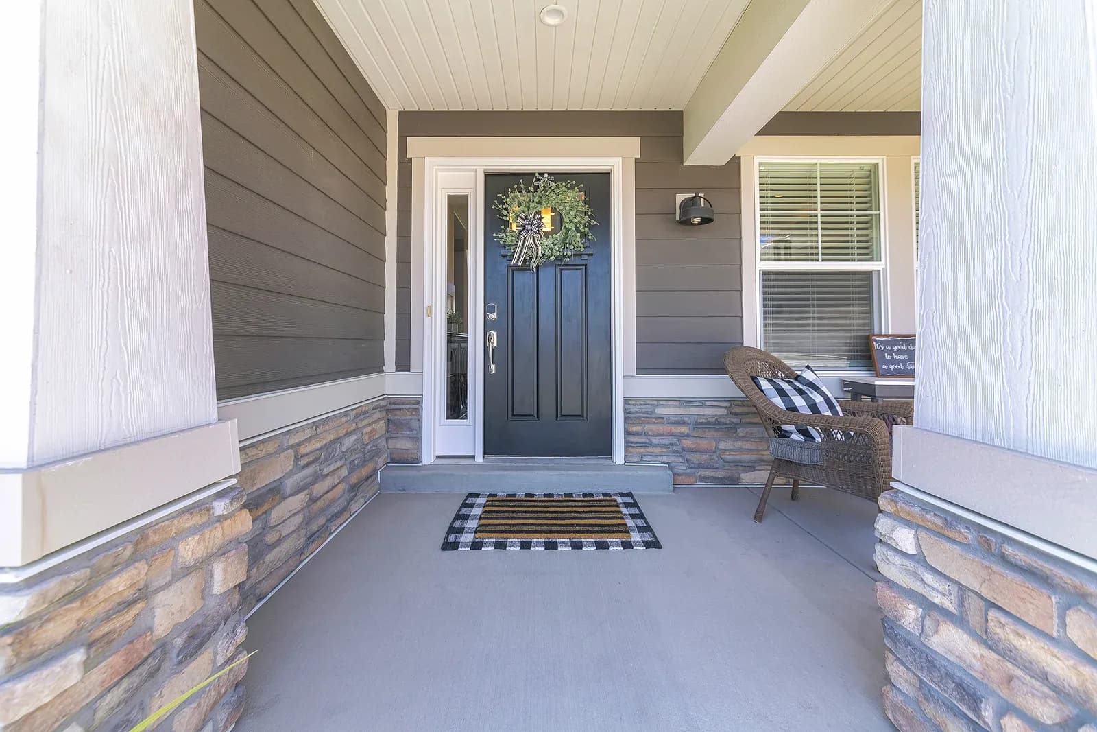 A photo from the porch facing the front door of a house. The door is black, has a narrow vertical window along the left-hand side, and has a wreath hanging on the front.