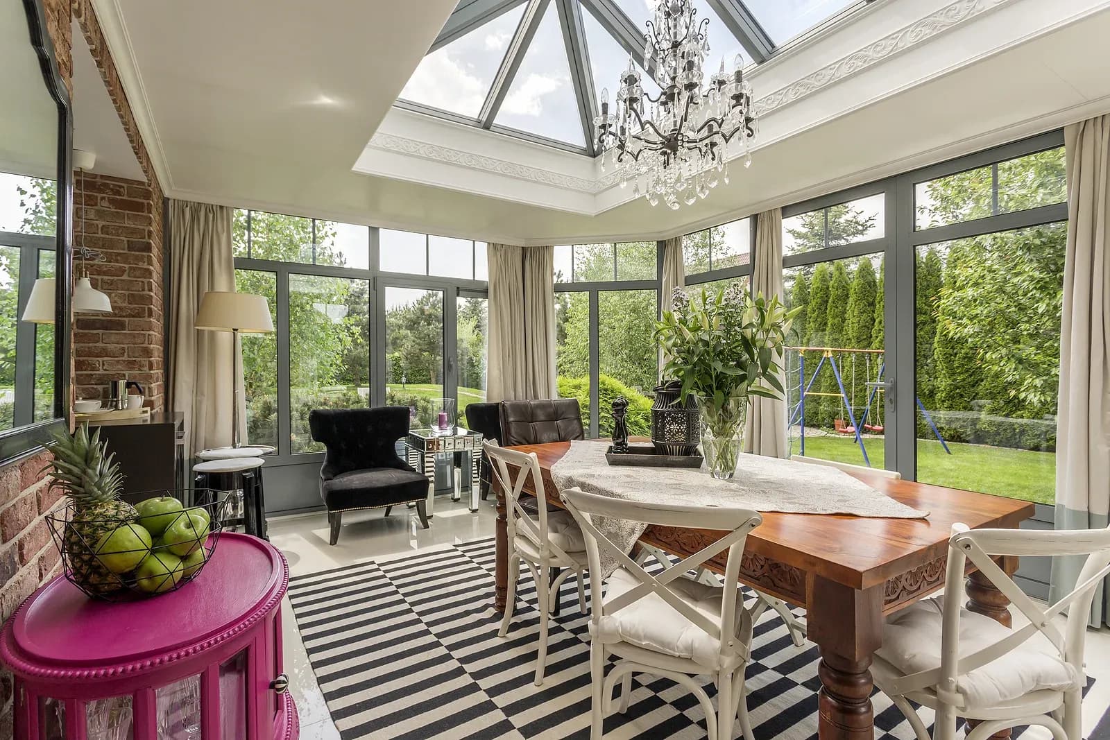 A photo of a dining table in the center of a sunroom. The walls and ceiling are all glass and look out into a backyard.