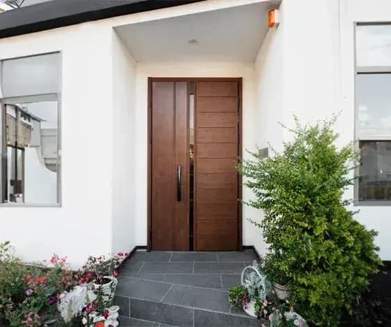 Modern white exterior entrance with dark wood double doors featuring vertical panels and handles, flanked by large windows under an overhanging roof, potted flowers and bush on stone steps.