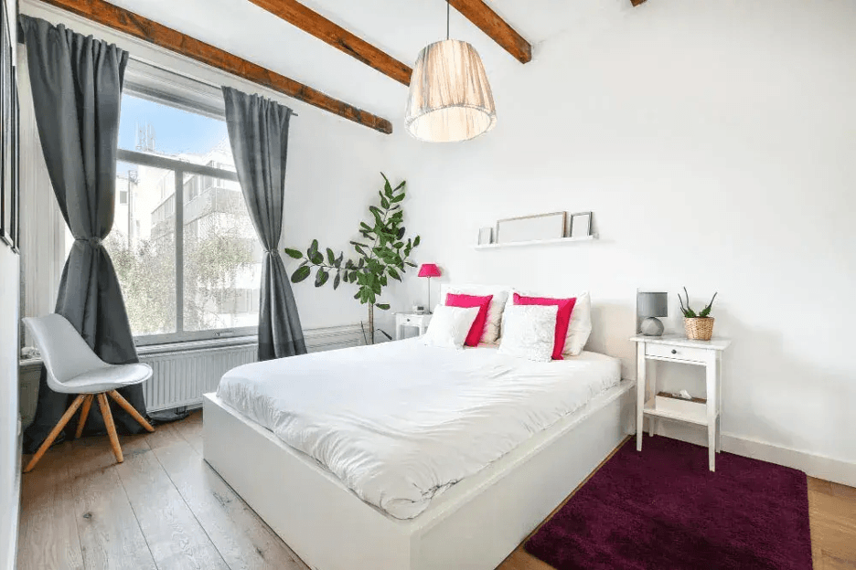 A modern minimalist bedroom with white walls and exposed wooden ceiling beams. The room includes a spacious bed, dark curtains, warm lighting, and decorative details that create an elegant and relaxing ambiance.