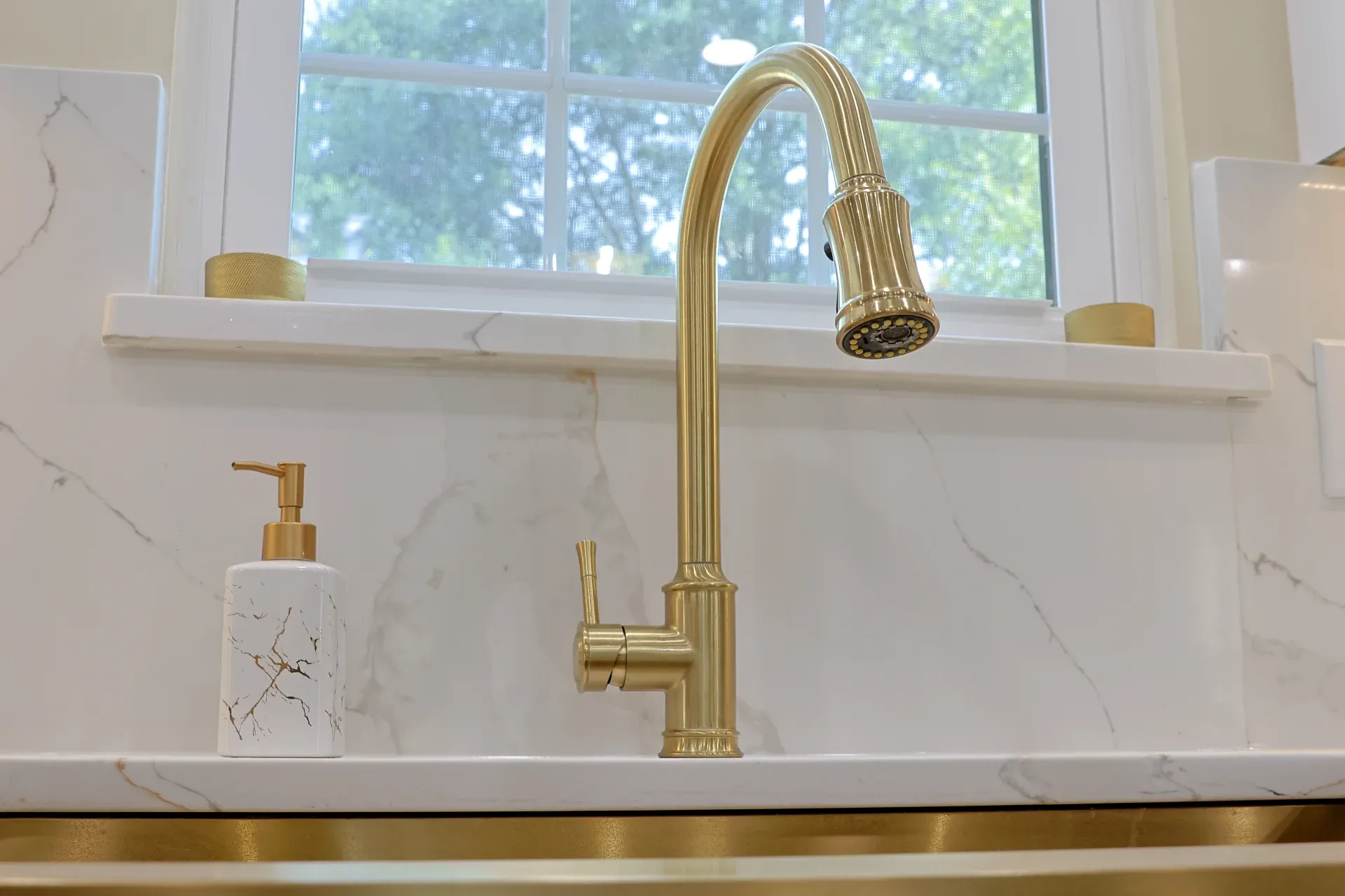 A gold faucet over a kitchen sink with white marbled backsplash and a matching soap pump.