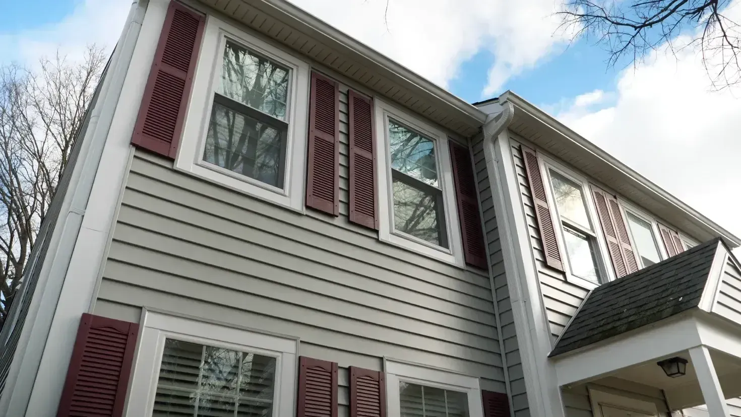 Low-angle view of a two-story residential home with light gray horizontal siding, white window trim, and dark red decorative shutters under a blue sky with light clouds.