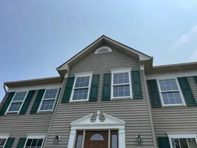 Two-story gray shingled house with white trim and dark green shutters, featuring a brown front door with sidelights, flanked by lanterns under a clear blue sky.