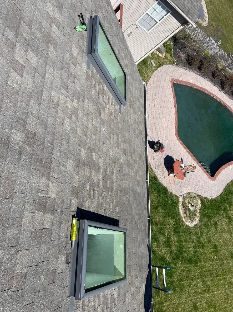 Aerial view of gray shingled rooftop with two skylights overlooking a curved green inground pool surrounded by brick pavers, lounge chairs, umbrellas, and green lawn.