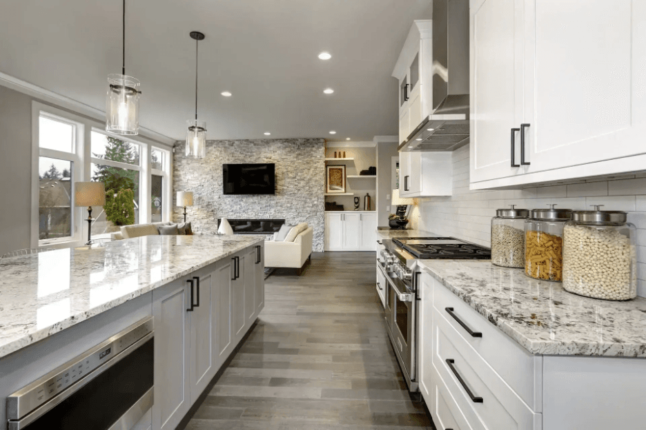 A spacious modern kitchen with white cabinets, marble countertops, and stainless steel appliances. The open-concept design, large windows, and soft lighting create a bright, clean, and sophisticated atmosphere.