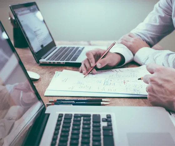 A person writes notes on a sheet of paper between two laptops on a desk.