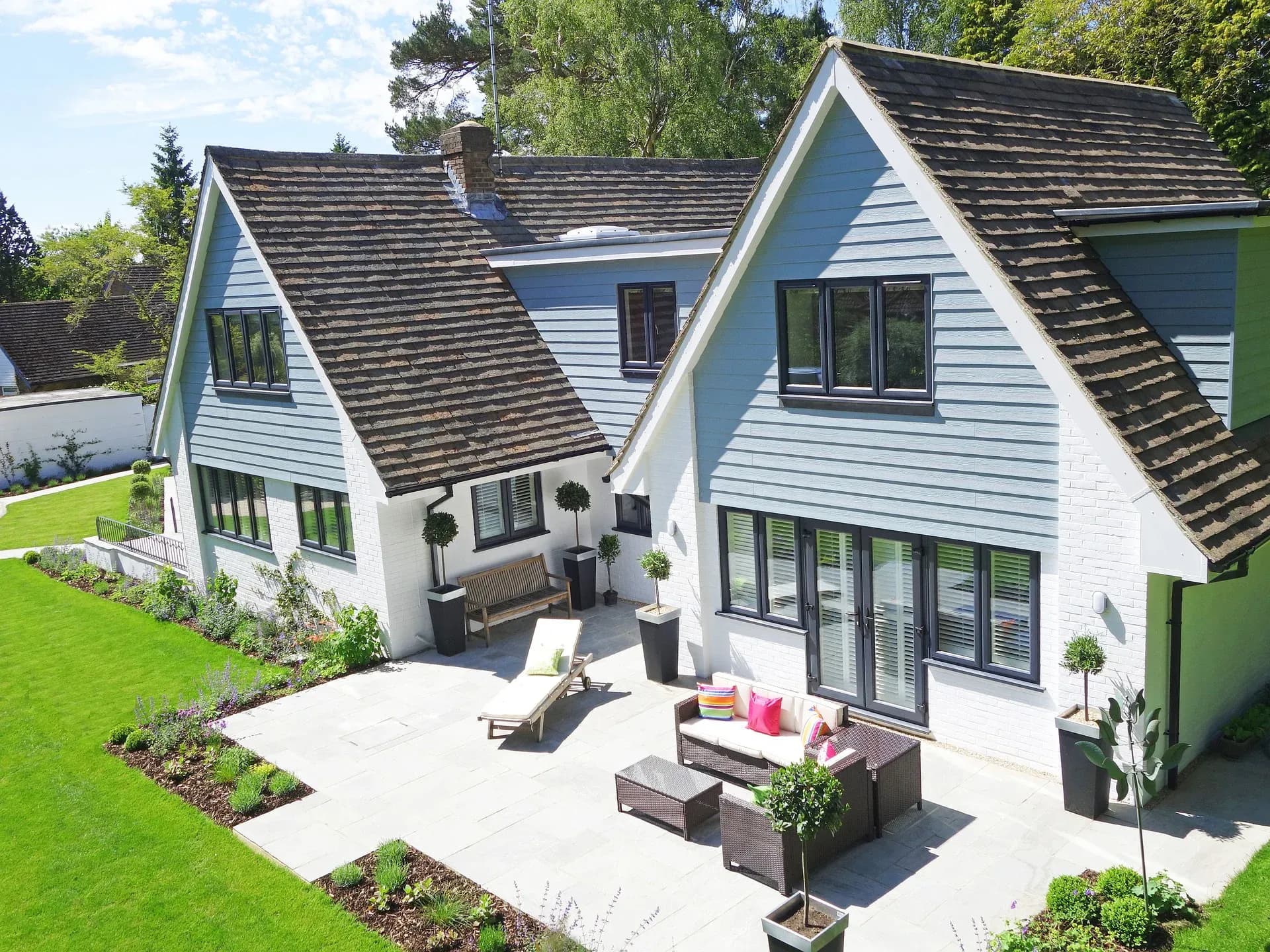 A photo of a house with white brick and gray-blue siding, black windows and doors, a large patio with lounge chairs and a small table.
