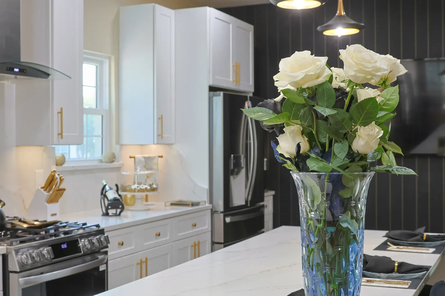 A vase of white roses sits on a white marble topped island. In the background, a stove, white and gold cabinets, and refrigerator are visible.