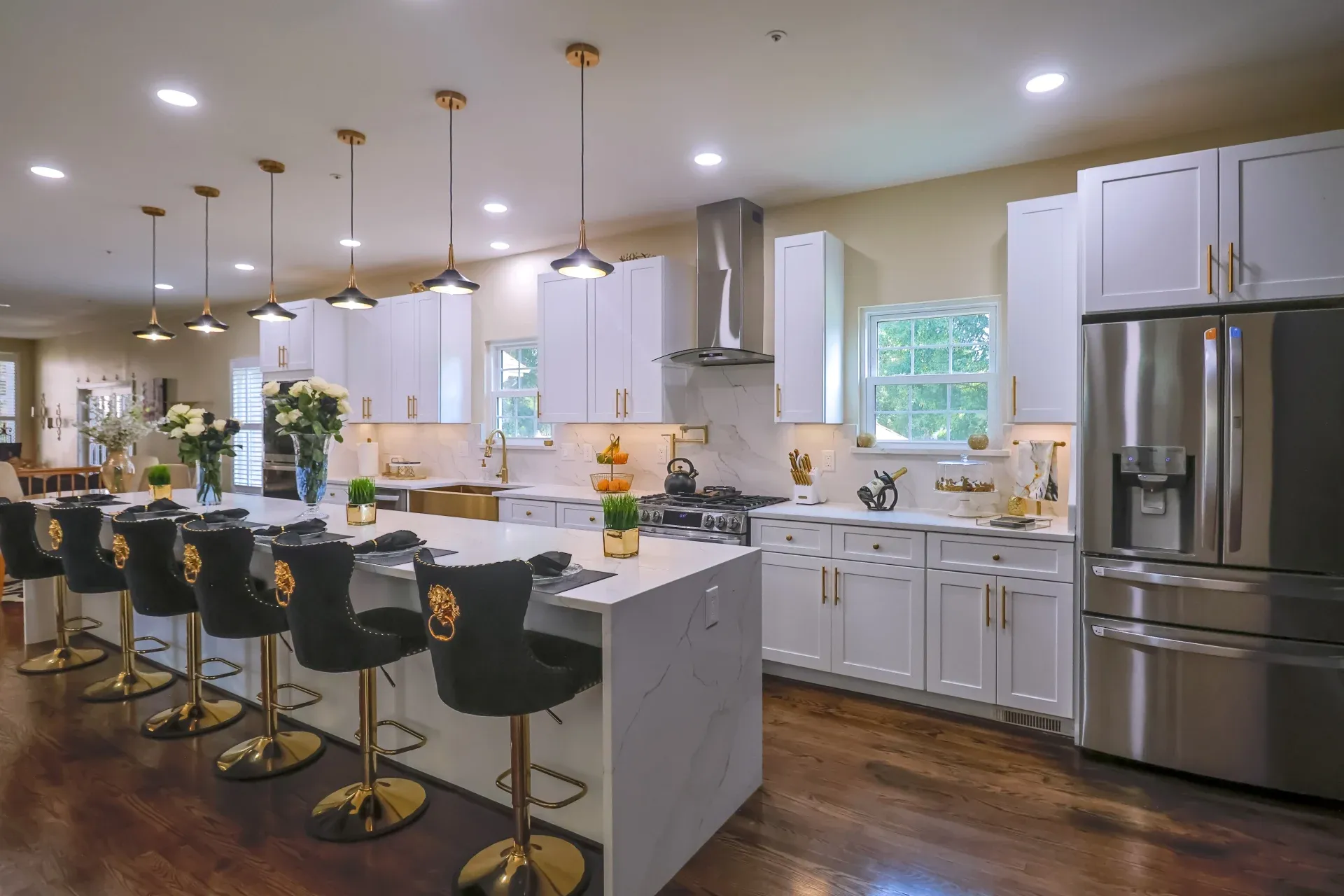 A wide view of a kitchen with a large island with black and gold bar stools around it, stainless steel appliances, and white cabinets with gold fixtures.