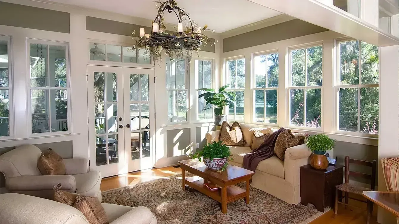 Bright sunroom with large windows, a cream-colored sofa with brown pillows, and a rustic branch chandelier. The room features a patterned area rug, a wooden coffee table with a potted plant, and white French doors.