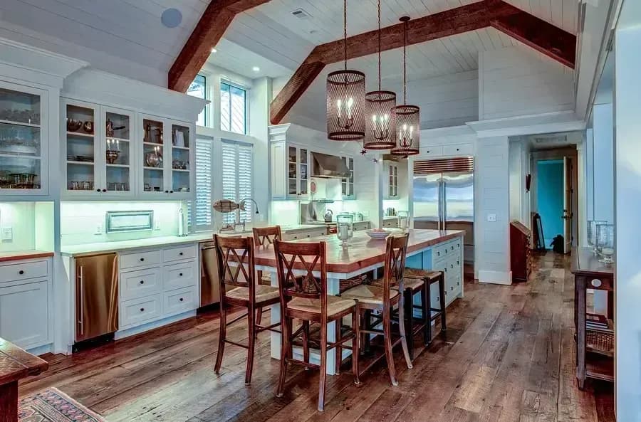 Modern farmhouse kitchen featuring white cabinetry, large wooden ceiling beams, a central island with a wood countertop, and three industrial mesh pendant lights over wooden barstools.