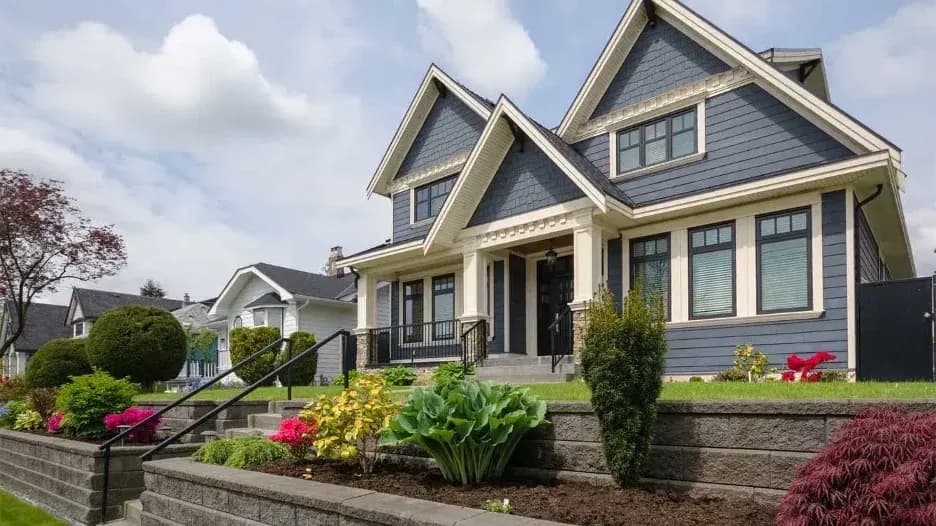 Modern suburban house with blue siding, white trim, and a landscaped front yard with flowers and shrubs on a raised garden bed.