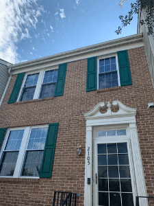 Red brick colonial house with white trim, green shutters, and front entrance door viewed from street level on clear day