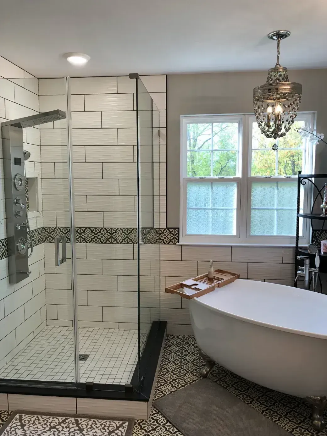 A spacious modern bathroom with a glass-enclosed shower featuring chrome rainfall showerhead and controls, white subway tile walls accented by Moroccan-patterned black trim. A large clawfoot bathtub sits nearby, under a sparkling crystal chandelier, with windows overlooking greenery, wooden tray, and gray rug on tiled floor.