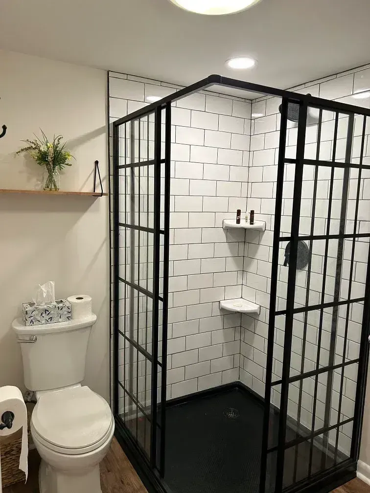 A modern bathroom featuring a walk-in shower with black metal frame and frosted glass panels, surrounded by white subway tile walls. It includes a white toilet, toilet paper holder, floating wooden shelf with fresh flowers and soap dispensers, recessed ceiling lights, and dark wood flooring