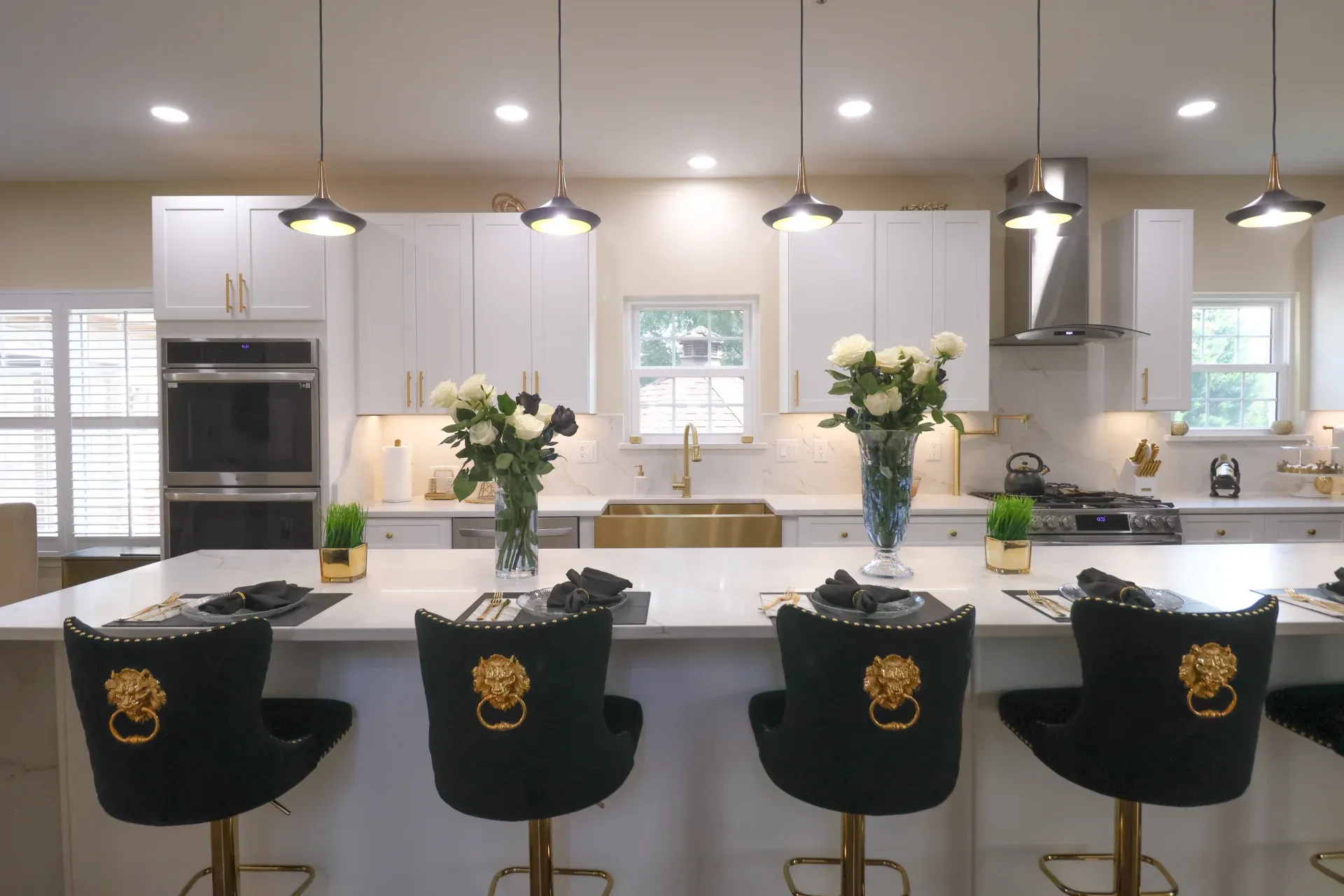 A row of black bar stools with gold lion heads face a kitchen island. There is a double oven towards the back left, and the kitchen has white cabinets and countertops. A brass-colored sink is visible in the center background.