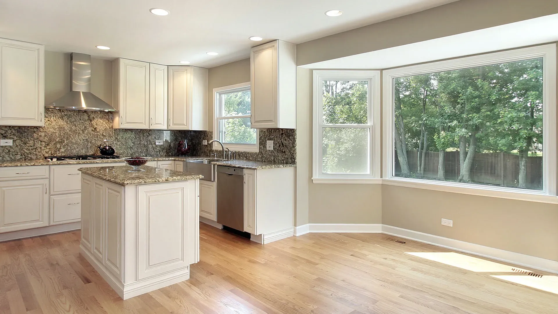 A kitchen with gray marbled countertops and backsplash, white cabinets, and stainless steel appliances. A large bay picture window is to the right.