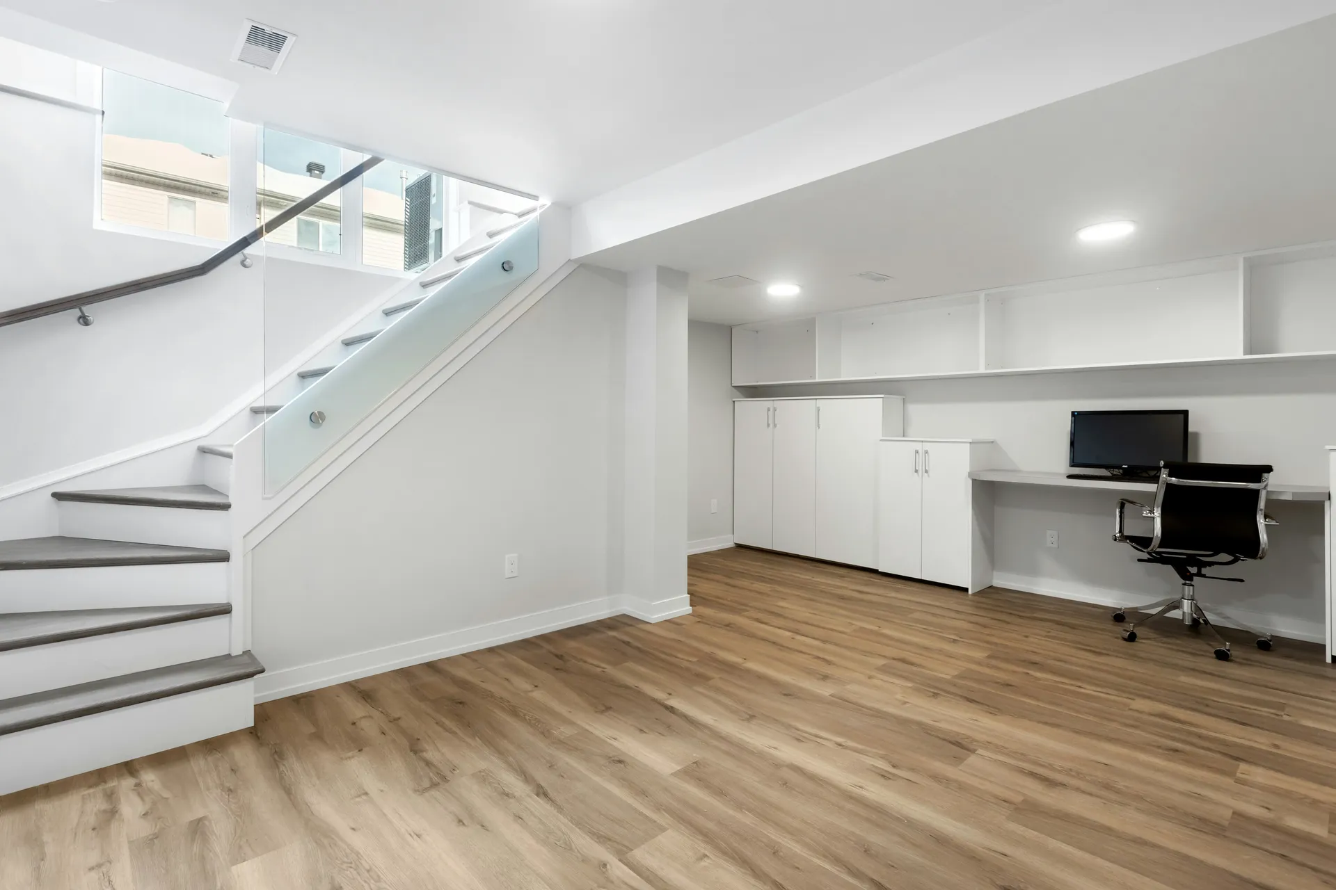 Finished basement remodel featuring luxury vinyl plank flooring, built-in white cabinetry, recessed lighting, glass stair railing, and a functional home office workspace.