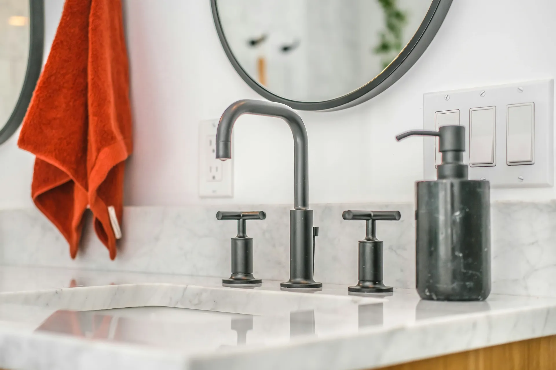 Modern bathroom vanity with marble countertop, matte black widespread faucet, round mirror, and warm accent towel, showcasing a clean, contemporary bathroom remodel.
