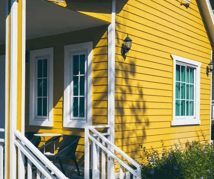 A cozy modern-style house with a bright yellow exterior and white accents. It features large windows, classic outdoor lighting, and a small porch with white railings that create a warm and peaceful atmosphere.