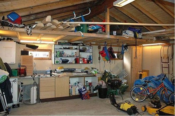 A wide, cluttered garage interior with unorganized open shelving, a messy wooden workbench, a chaotic loft space with stored wood, and scattered items like a bicycle, power tools, and bags